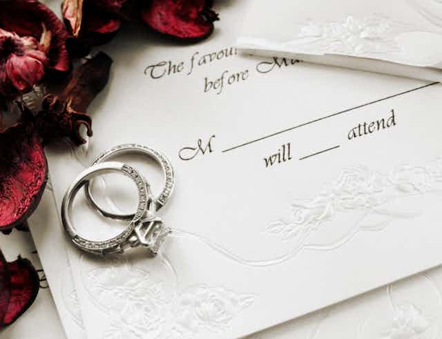 A wedding invitation card with a request to RSVP sits on a table with a pair of rings and some dried flowers