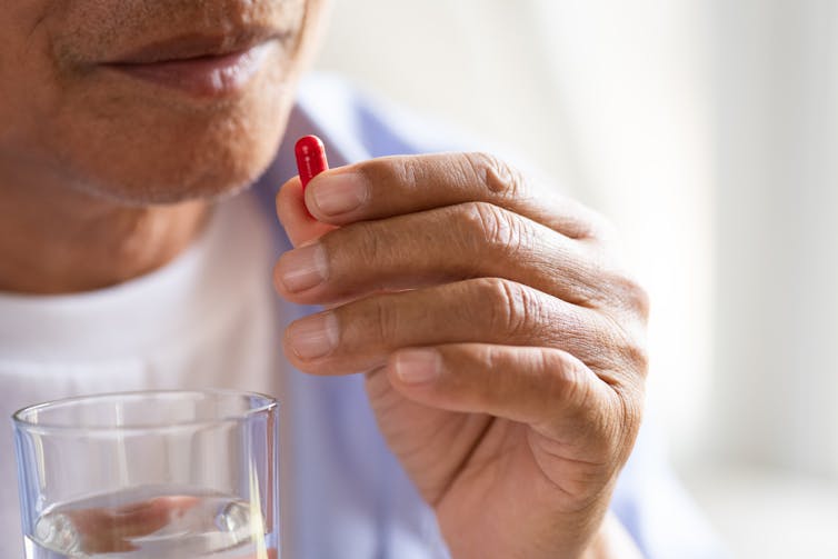Older man holds pill to his mouth in one hand and a glass of water in the other.
