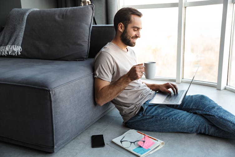 A man sits on the floor on his laptop, with a cup of coffee in hand.