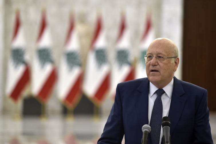 Old man wearing a suit speaks into microphones in front of a row of Lebanese flags.