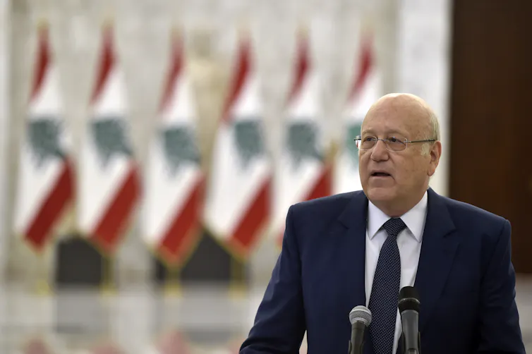 Old man wearing a suit speaks into microphones in front of a row of Lebanese flags.