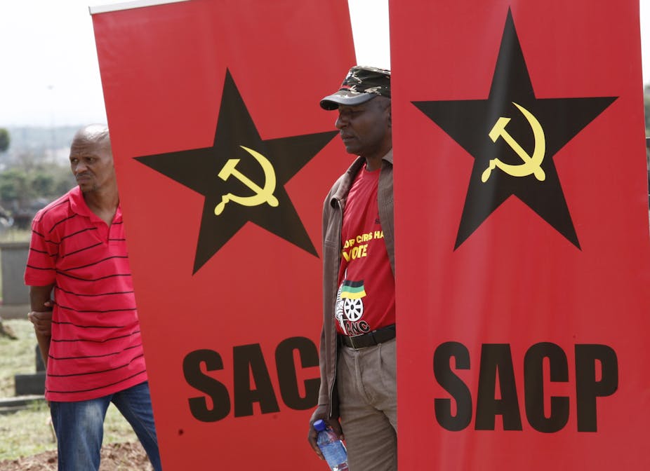 A wearing an African National Congress T.shirt stands between two South African Communist Party banners showing the hammer and circle.