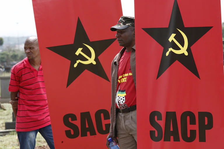A wearing an African National Congress T.shirt stands between two South African Communist Party banners showing the hammer and circle.