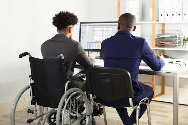 Two men are seen at an office computer, one of them in a wheelchair.