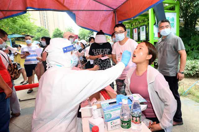 A young Chinese woman receives a COVID test from someone in full protective clothing while people wait in line.