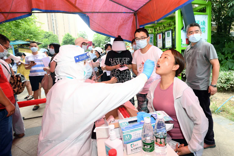 A young Chinese woman receives a COVID test from someone in full protective clothing while people wait in line.
