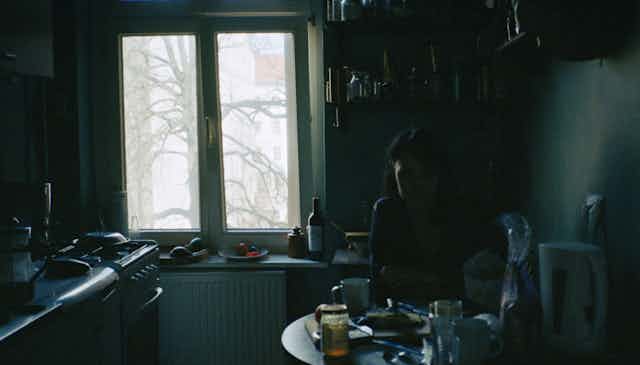 A woman sits alone in a dark kitchen, staring at the table and not touching her food.