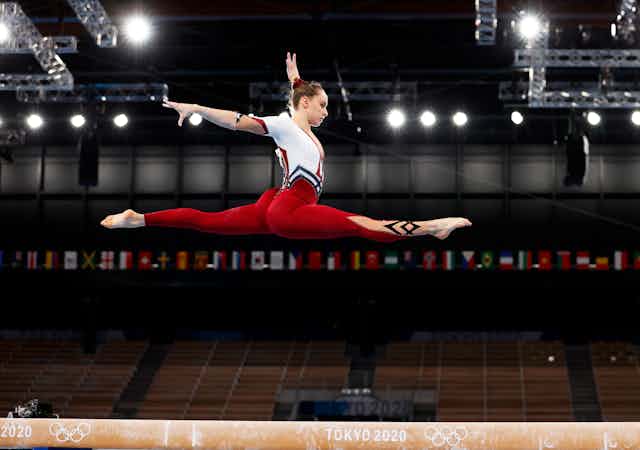 German gymnast Sarah Voss leaping high above the beam in a full-body red and white unitard rather than a revealing leotard.