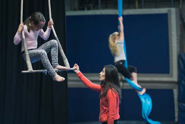 Une fille sur un trapèze recevant des instructions, avec une autre fille grimpant à une corde en arrière-plan.