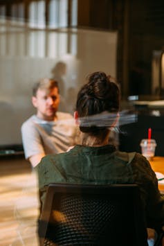 A man and woman talk in an office setting, the woman with her back to the camera.