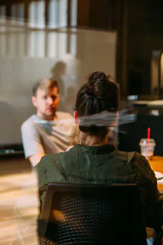 A man and woman talk in an office setting, the woman with her back to the camera.