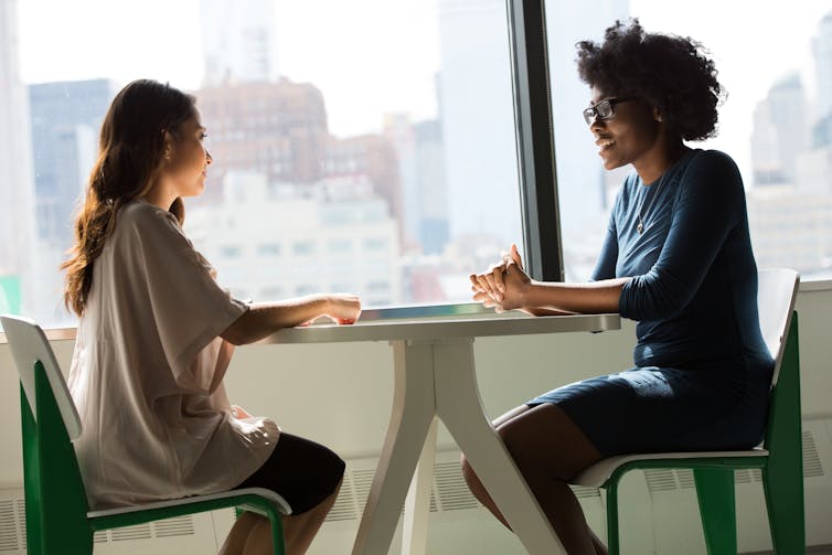 Two women sit talking at a table in an office setting.