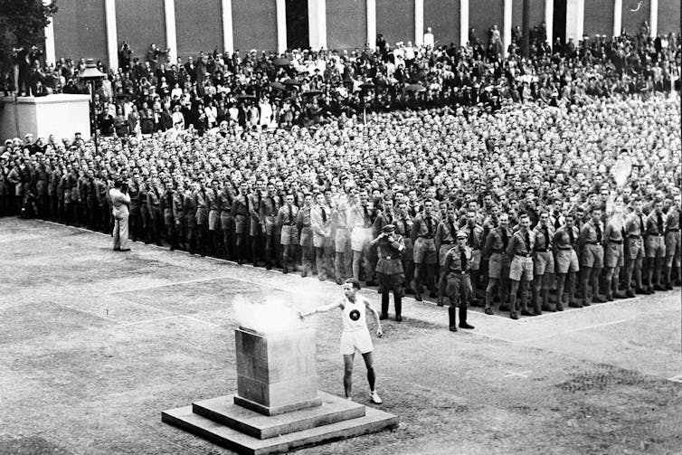 Man lighting Olympic torch in foreground with lines of Nazi soliders lined up behind him.