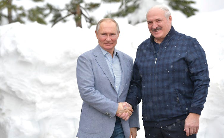 Putin and Lukashenko shake hands while both smiling and facing the camera. They are outdoors against a snowy background.