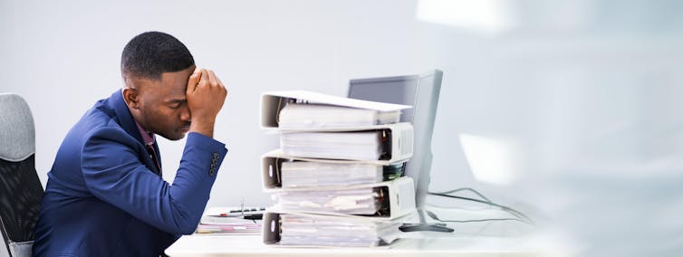An office worker sits in front of a desk piled high with files, holding his head in his hand