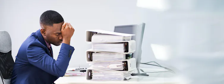 An office worker sits in front of a desk piled high with files, holding his head in his hand