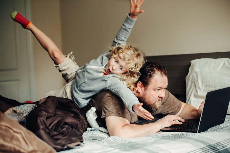 A father works at a laptop as his daughter jumps on his back