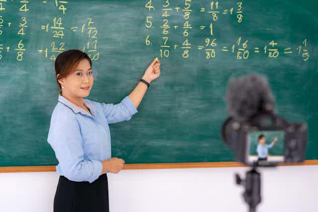 A teacher points to a blackboard as she films a lesson for her pupils working at home