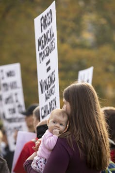 A woman holding a baby holds a placard reading 'Forced medical procedures equals violence against children.'