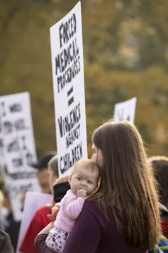 A woman holding a baby holds a placard reading 'Forced medical procedures equals violence against children.'