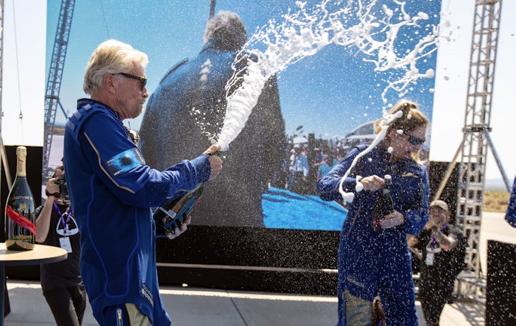 Richard Branson in a blue jumpsuit celebrated his successful sub-orbital flight to space by spraying champagne on a stage.