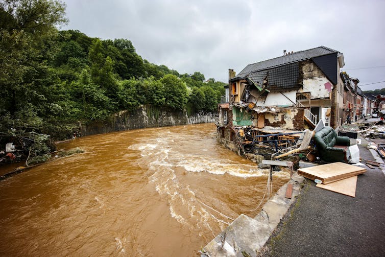 Muddy water water pours past a home where the side has been ripped open revealing the interior of rooms up to the second floor