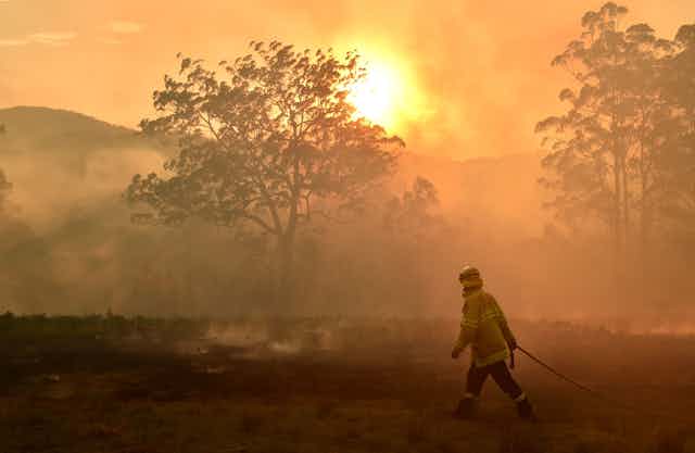 A wildland firefighter with a hose walks through a burned landscape with smoke filling the sky