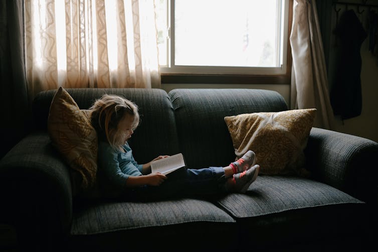 Young girl reading book on couch next to window