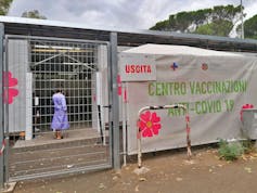 A woman wearing a purple hospital gown can be seen through a metal fence. A large colorful sign reads 'Centro Vaccinazioni Anti-Covid 19' in green letters
