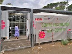 Une femme portant une chemise d'hôpital violette peut être vue à travers une clôture métallique.  Un grand panneau coloré indique 'Centro Vaccinazioni Anti-Covid 19' en lettres vertes