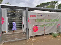 A woman wearing a purple hospital gown can be seen through a metal fence. A large colorful sign reads 'Centro Vaccinazioni Anti-Covid 19' in green letters