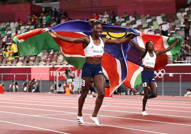 Two young omen run on an athletics track, each holding a Namibian flag behind them.