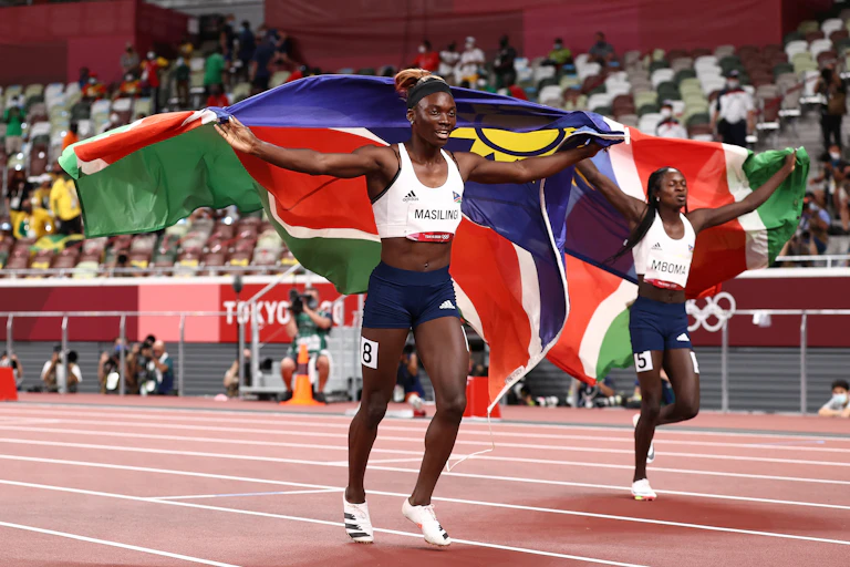Two young omen run on an athletics track, each holding a Namibian flag behind them.