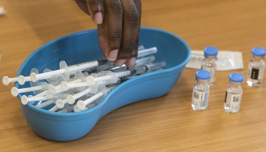 A hand reaches into a blue, kidney-shaped bowl full of syringes. There are a few vials of clear liquid with blue lids standing alongside it.