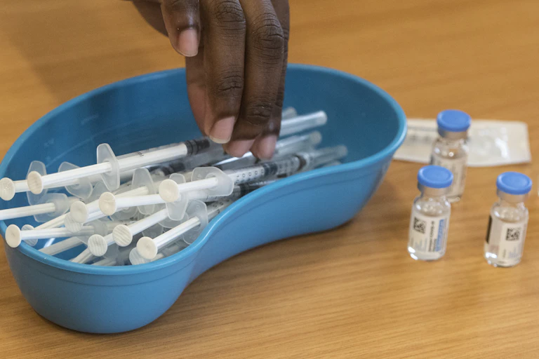 A hand reaches into a blue, kidney-shaped bowl full of syringes. There are a few vials of clear liquid with blue lids standing alongside it.