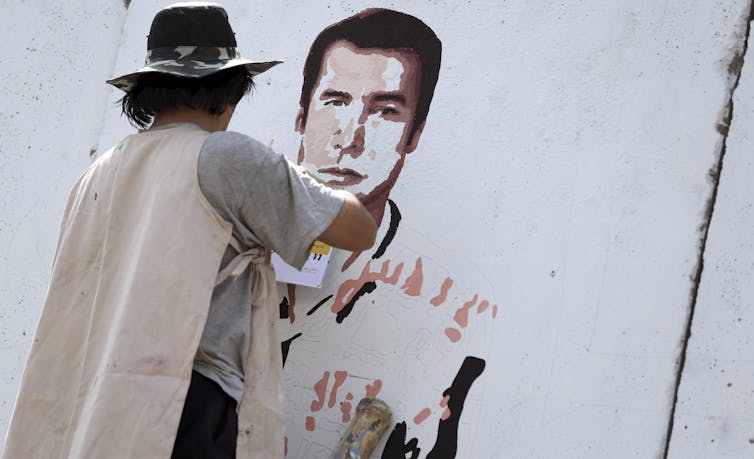 An artist wearing a hat paints the portrait of a man on a mural.