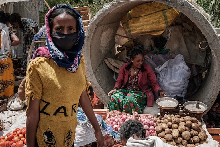Woman sits behind pile of vegetables as another woman walks past