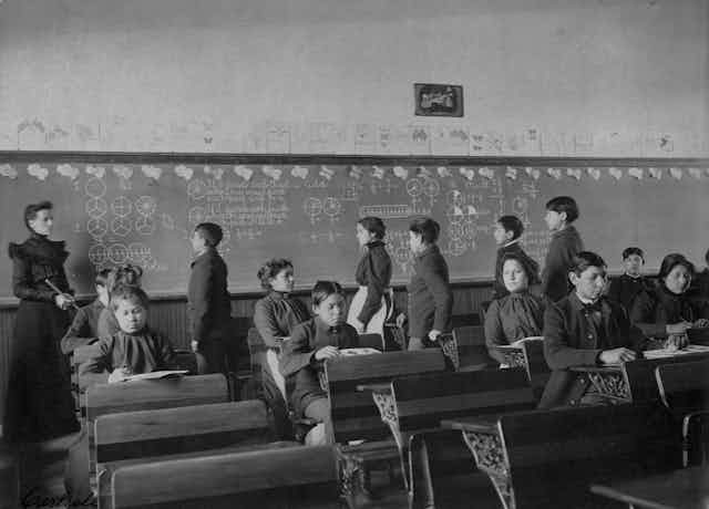 Archival photo of Native American students sitting at desks and lined up in front of chalkboard in classroom