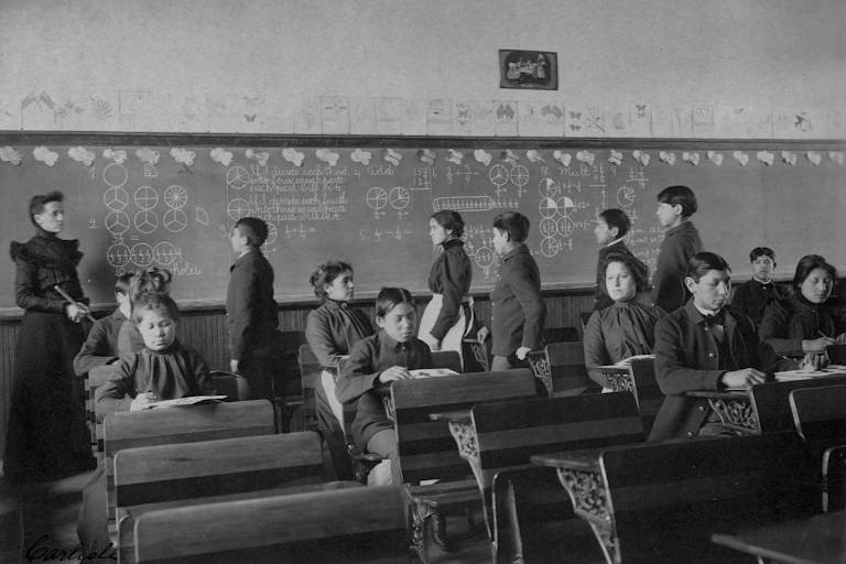 Archival photo of Native American students sitting at desks and lined up in front of chalkboard in classroom