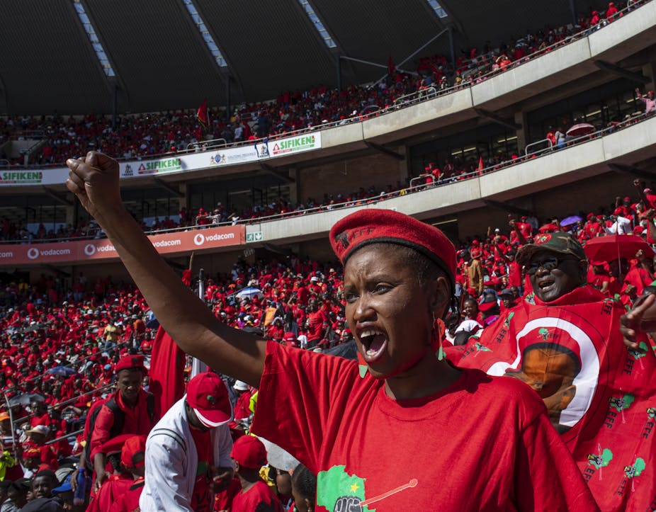 A woman raises her right hand in a black power salute. She is dressed in a red T-shirt and beret and is surrounded by similarly dressed people in a stadium.