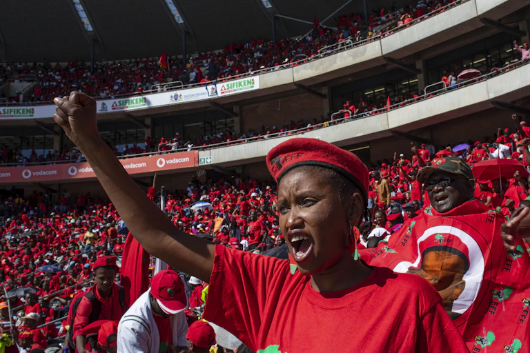 A woman raises her right hand in a black power salute. She is dressed in a red T-shirt and beret and is surrounded by similarly dressed people in a stadium.