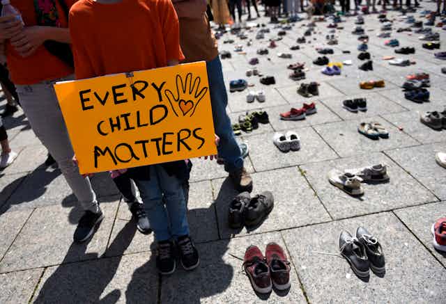 A child wearing an orange shirt holds a sign that reads 'every child matters' they are surrounded by hundreds of childrens shoes