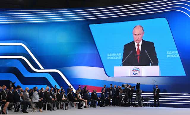 Putin's face is project on a large screen; people sit in chairs, wearing face masks, watching the screen in a large convention hall