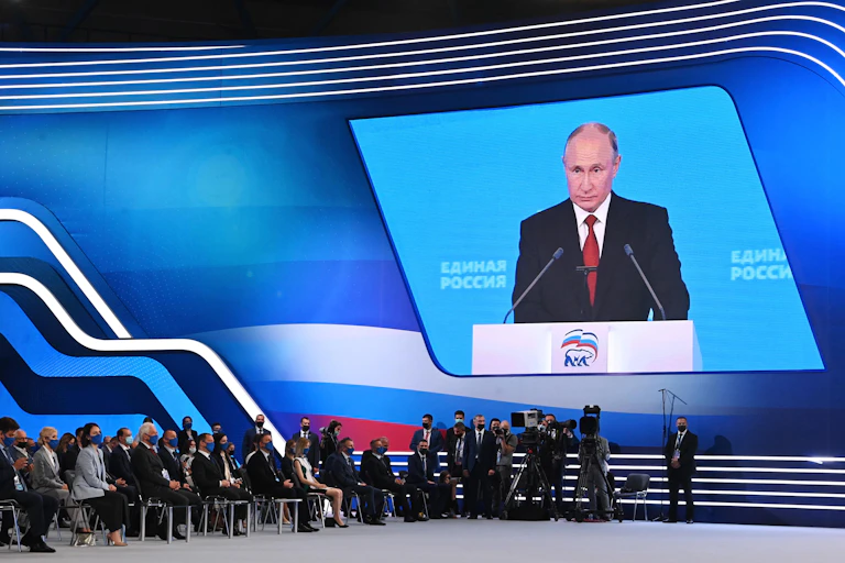 Putin's face is project on a large screen; people sit in chairs, wearing face masks, watching the screen in a large convention hall
