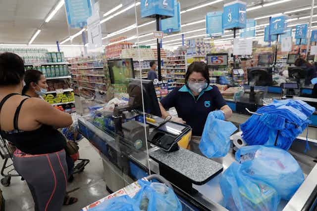 A cashier in a grocer store wearing a mask bags grocers as two customers look on