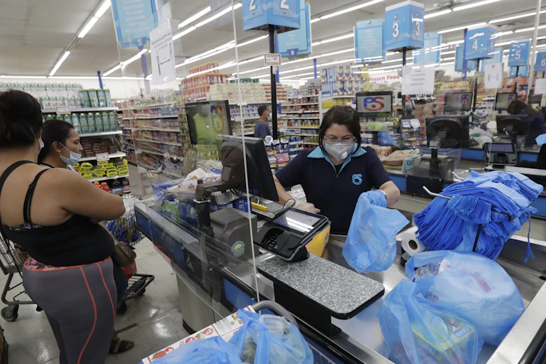 A cashier in a grocer store wearing a mask bags grocers as two customers look on