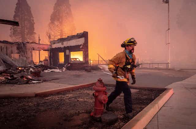 A firefighter in front of a destroyed building, smoke haze and fire in the background