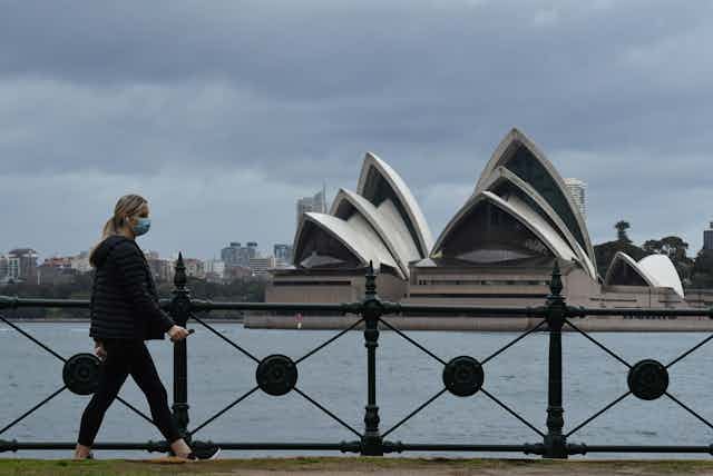 Woman wearing mask walks in front of Sydney Opera House