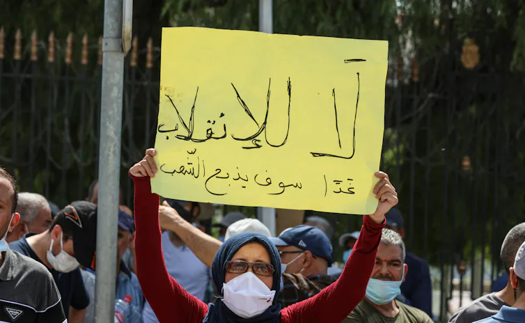 A Tunisian protester wearing a COVID mask holds up a sign reading 'Coup d'etat' in Arabic script.