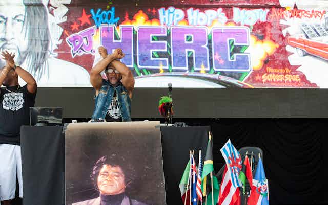 A rapper holds up both of his arms crossed while performing on stage. Behind him is a  giant billboard that reads 'D.J. HERC' in graffiti.